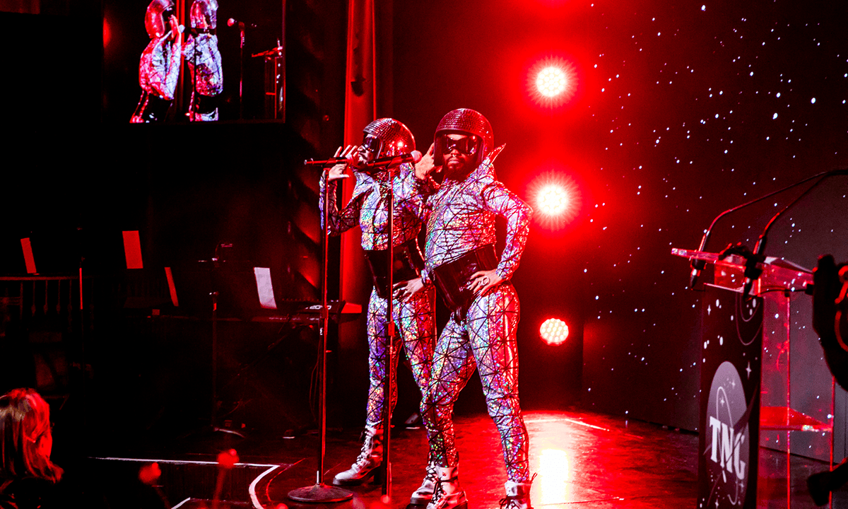 Two people stand at microphones wearing a sparkly suit and helmets.