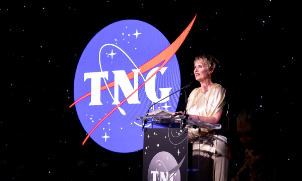 A woman stands at a podium talking to a crowd with the TNG logo hanging behind her.