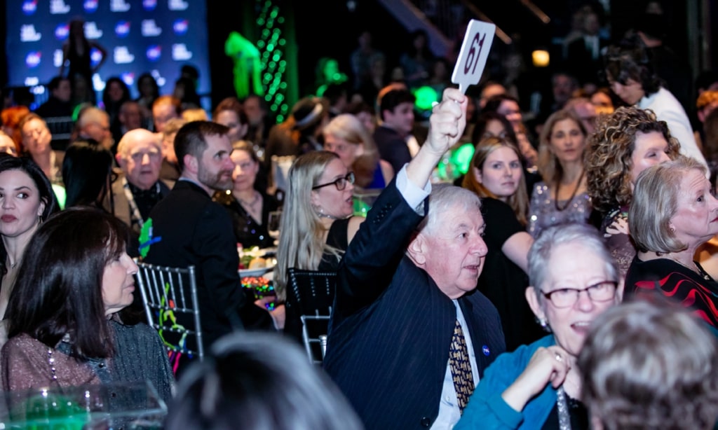 A room filled with people sitting at tables and one older man holding up a bidding number saying sixty one.