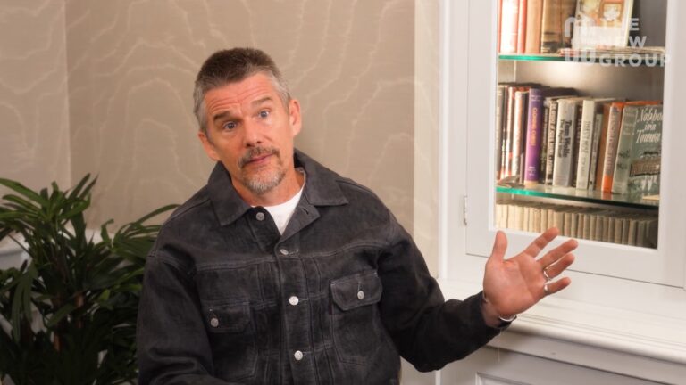 Ethan Hawke sitting beside a bookshelf with his hand up.