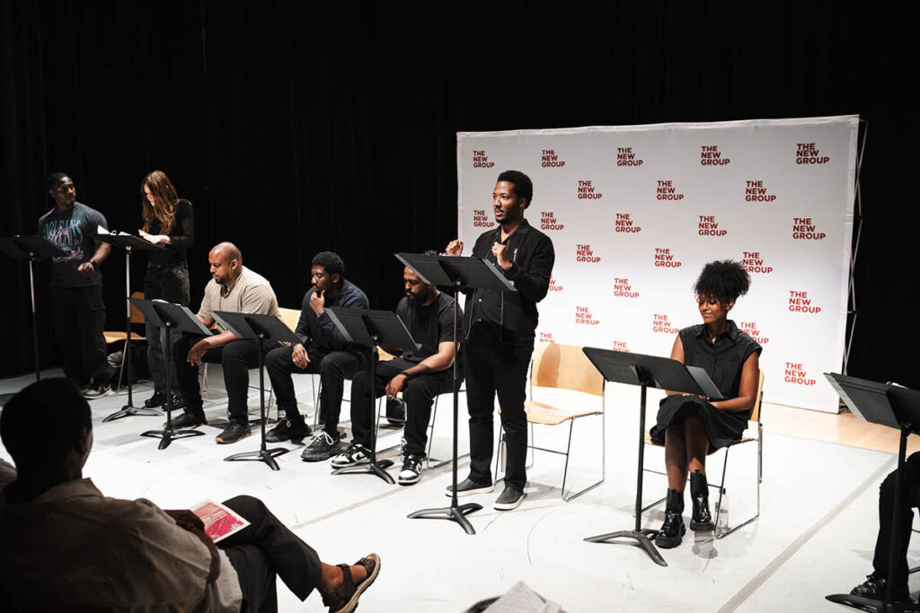 Actors sit in front of music stands with The New Group banner behind them