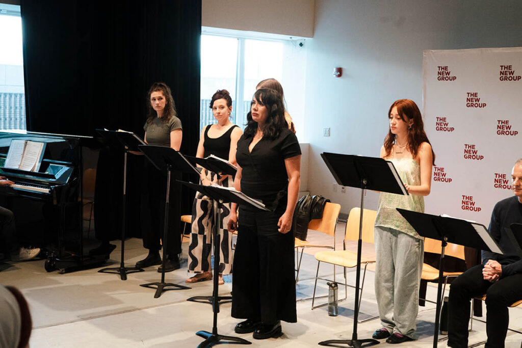 Actors sit in front of music stands with The New Group banner behind them