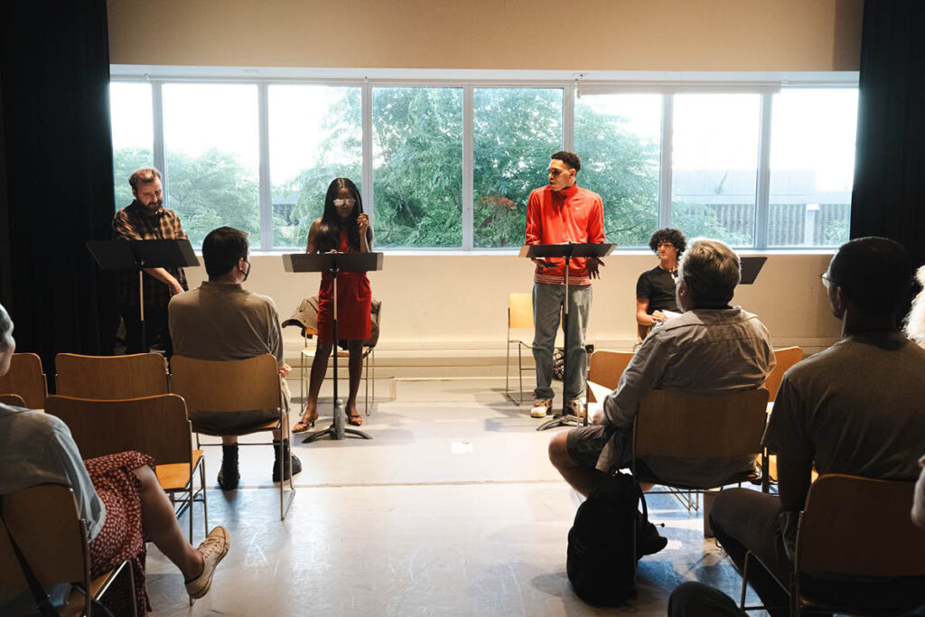 Two actors stand in front of music stands. A seated audience watches them.