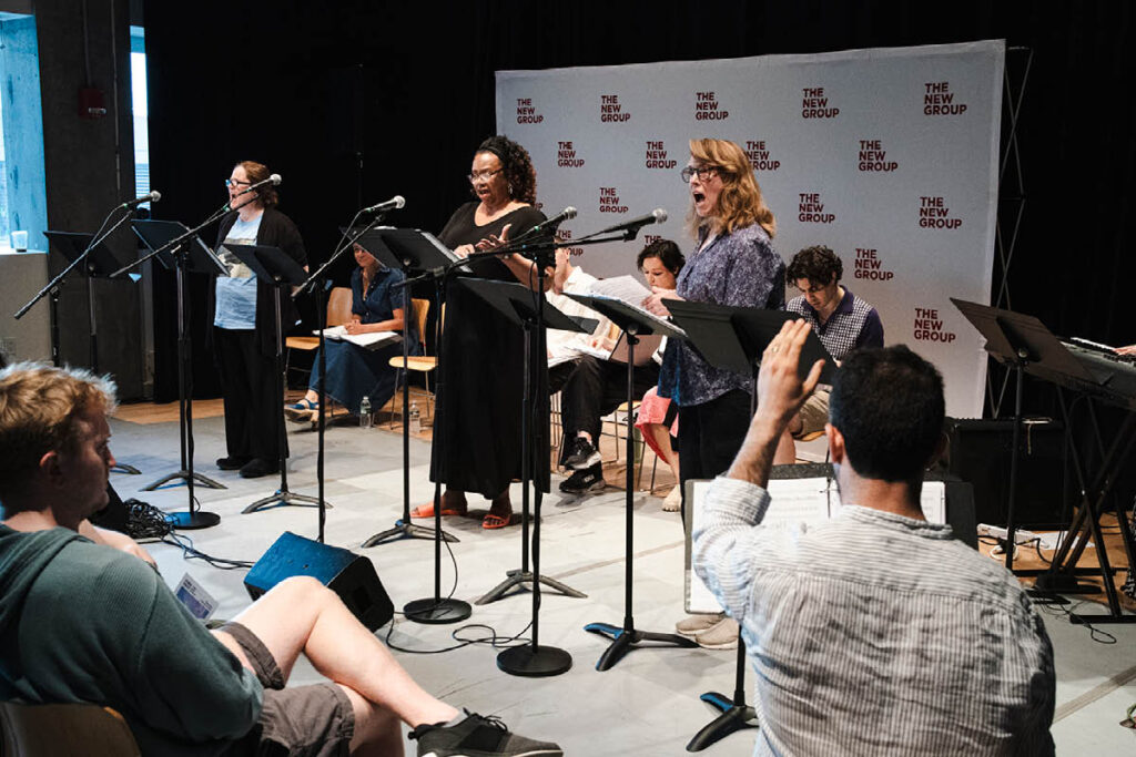 Actors sit in front of music stands with The New Group banner behind them